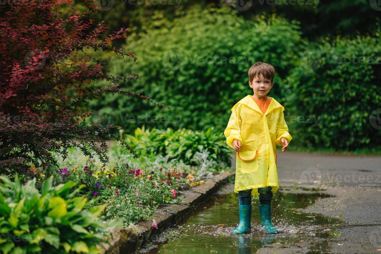 Little boy playing in rainy summer park. Child with umbrella, waterproof coat and boots jumping in puddle and mud in the rain. Kid walking in summer rain Outdoor fun by any weather. happy childhood. photo