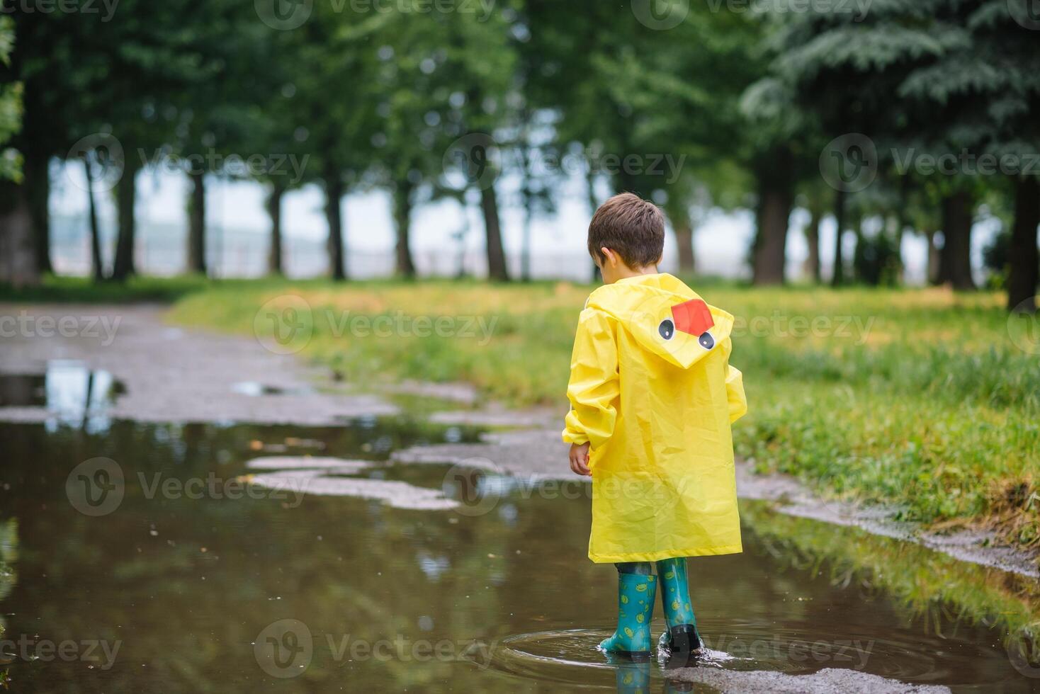 Little boy playing in rainy summer park. Child with umbrella, waterproof coat and boots jumping in puddle and mud in the rain. Kid walking in summer rain Outdoor fun by any weather. happy childhood photo