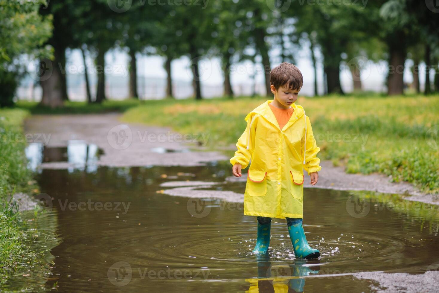 Little boy playing in rainy summer park. Child with umbrella, waterproof coat and boots jumping in puddle and mud in the rain. Kid walking in summer rain Outdoor fun by any weather. happy childhood photo