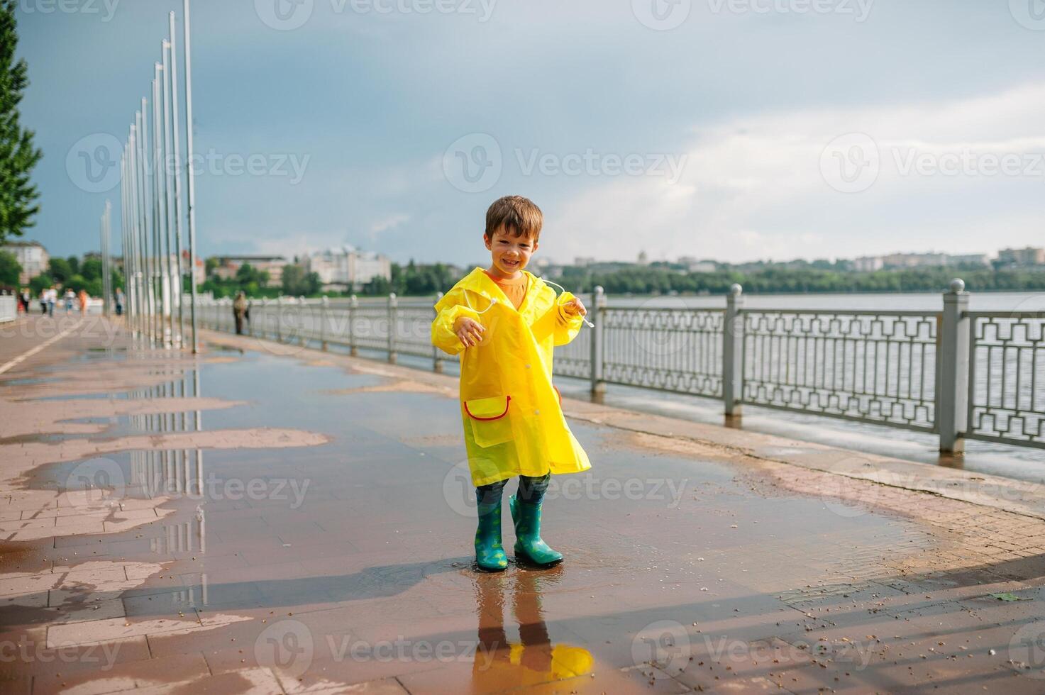 Little boy playing in rainy summer park. Child with umbrella, waterproof coat and boots jumping in puddle and mud in the rain. Kid walking in summer rain Outdoor fun by any weather. happy childhood. photo