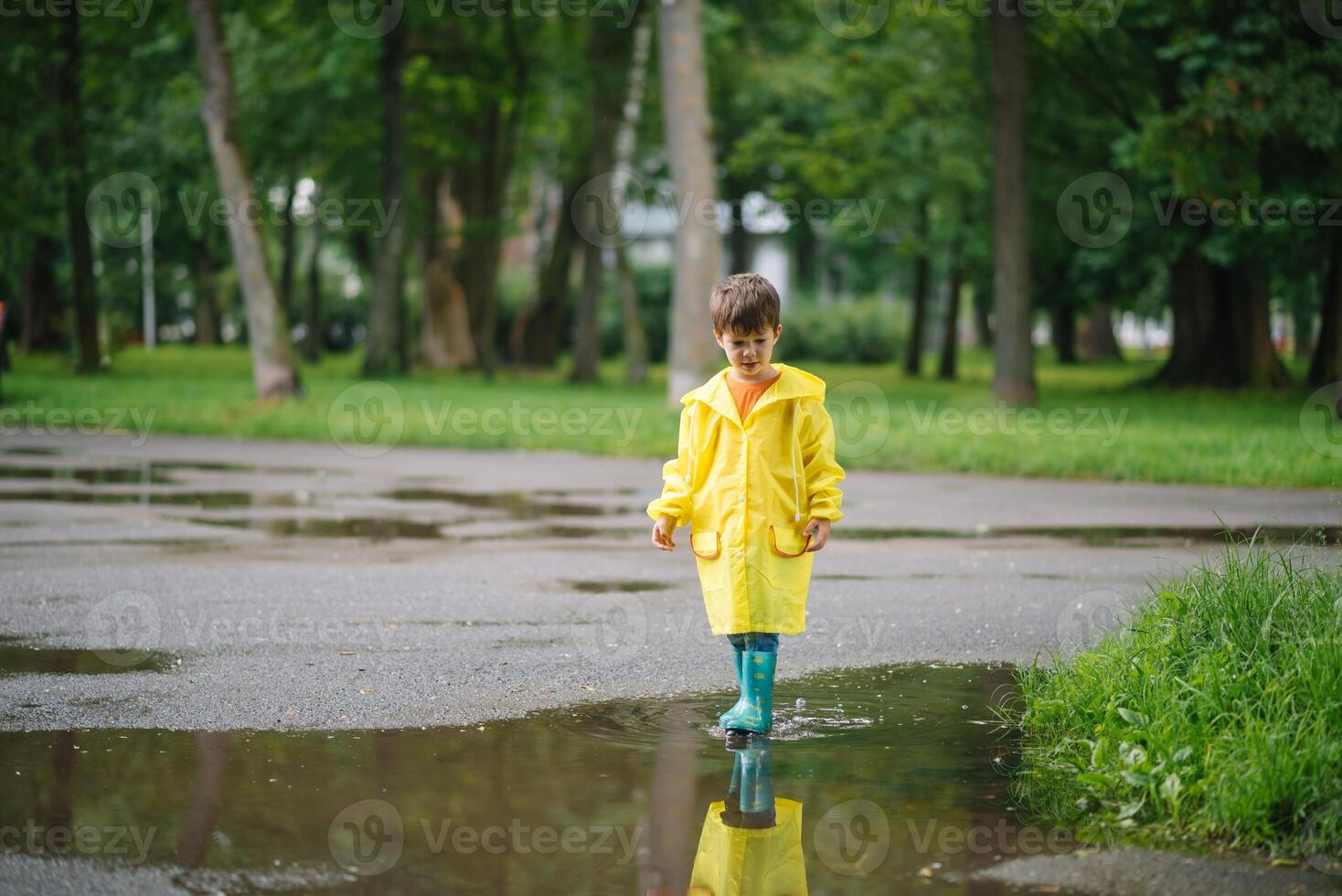 Little boy playing in rainy summer park. Child with umbrella, waterproof coat and boots jumping in puddle and mud in the rain. Kid walking in summer rain Outdoor fun by any weather. happy childhood photo