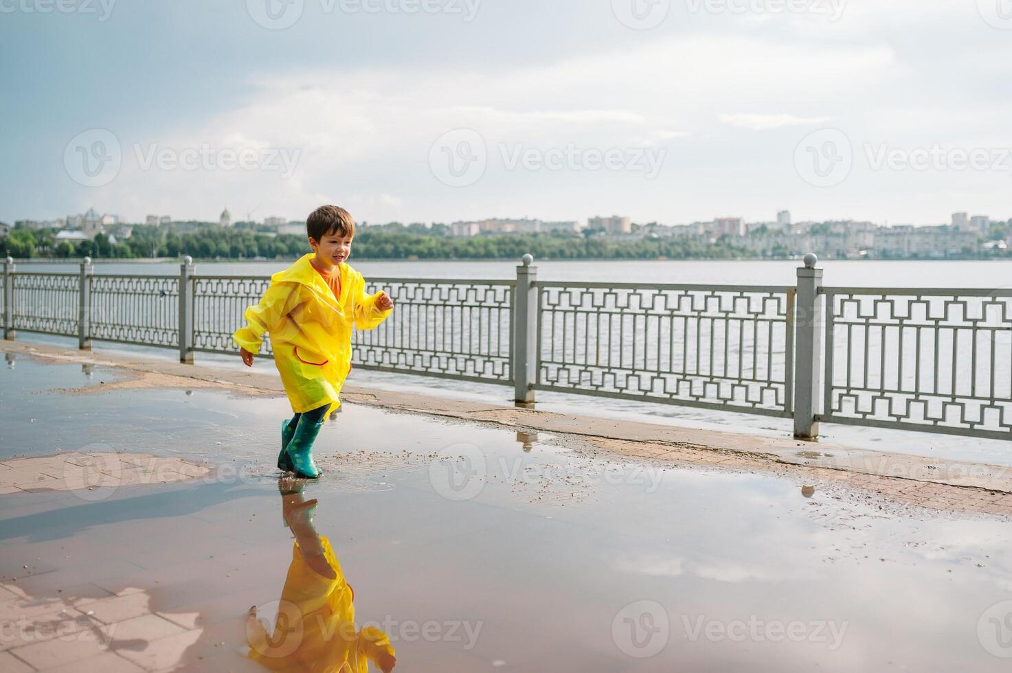 Little boy playing in rainy summer park. Child with umbrella, waterproof coat and boots jumping in puddle and mud in the rain. Kid walking in summer rain Outdoor fun by any weather. happy childhood. photo