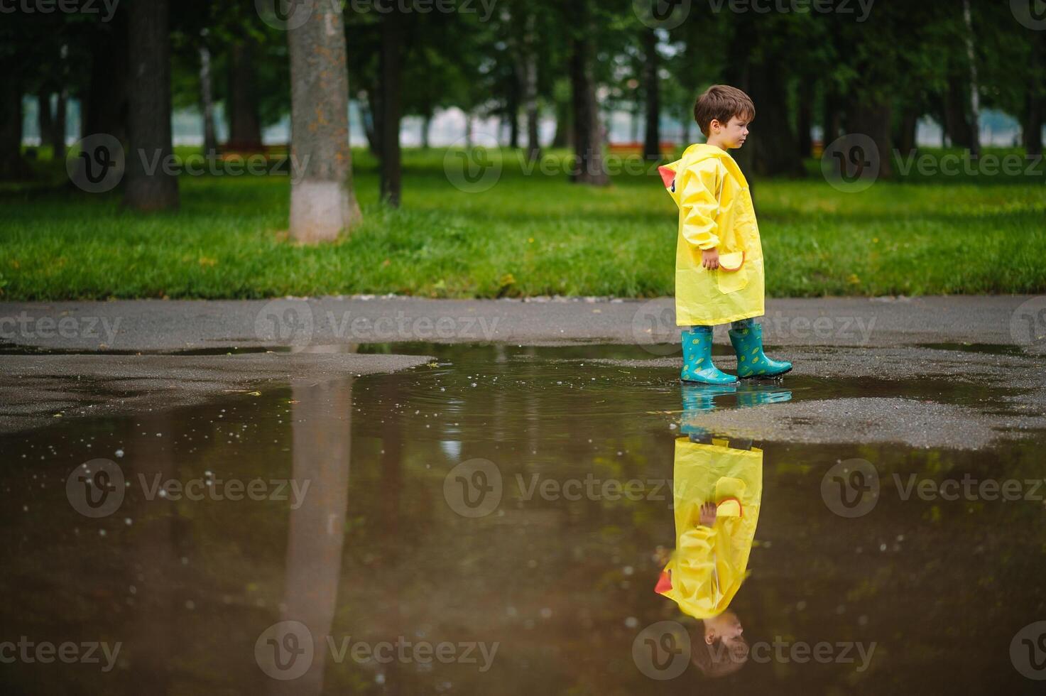 Little boy playing in rainy summer park. Child with umbrella, waterproof coat and boots jumping in puddle and mud in the rain. Kid walking in summer rain Outdoor fun by any weather. happy childhood. photo