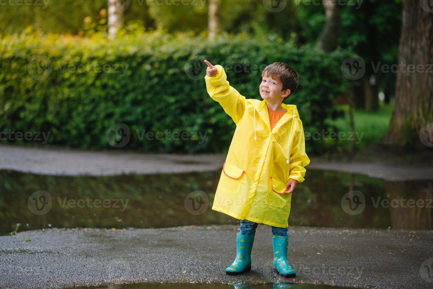 Little boy playing in rainy summer park. Child with umbrella, waterproof coat and boots jumping in puddle and mud in the rain. Kid walking in summer rain Outdoor fun by any weather. happy childhood photo