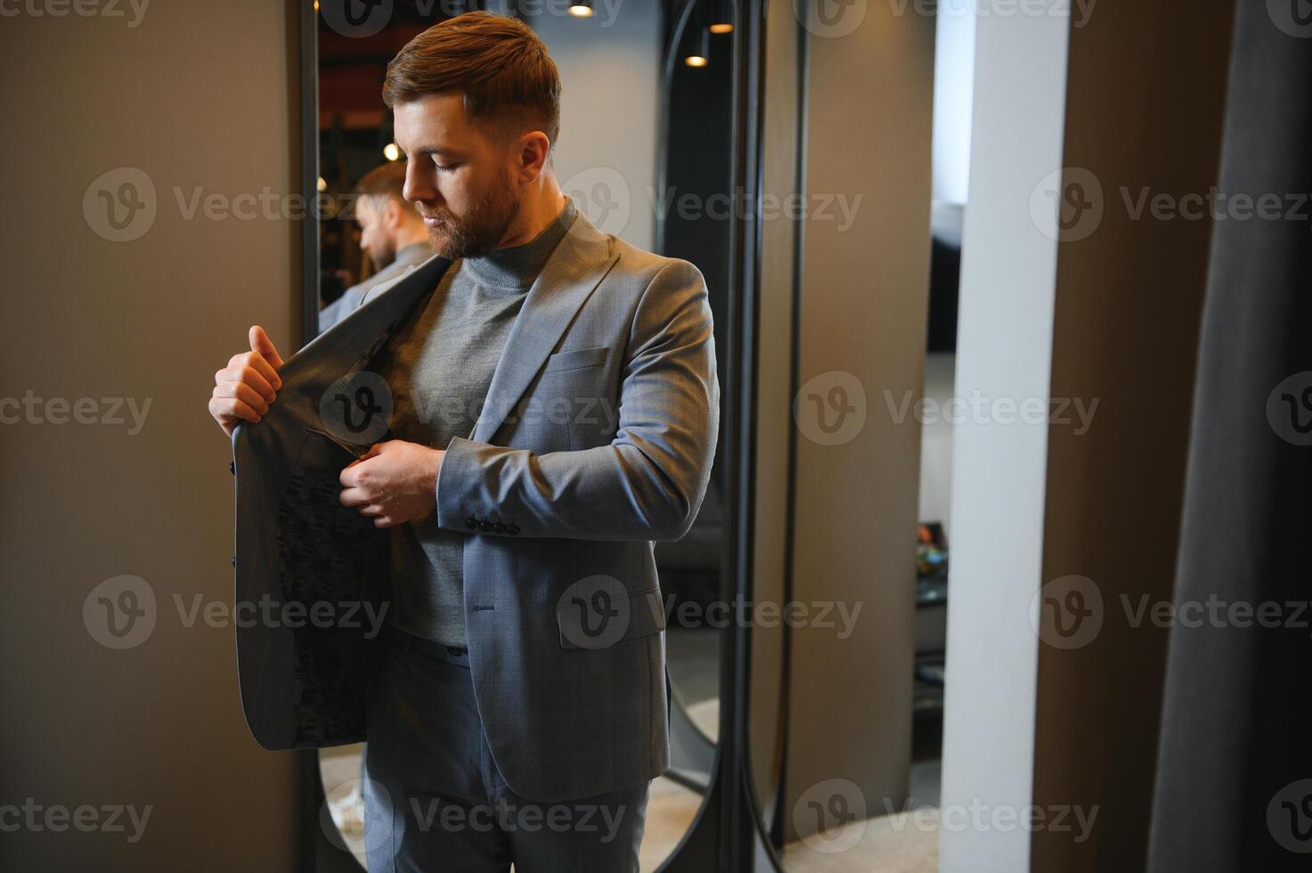 Modern young handsome businessman dressed in classical suit adjusting a tie in front of the mirror while standing in the suit shop photo