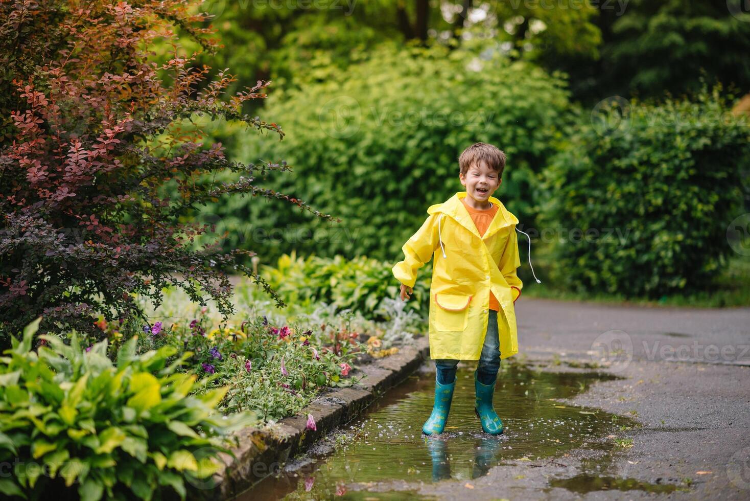 Little boy playing in rainy summer park. Child with umbrella, waterproof coat and boots jumping in puddle and mud in the rain. Kid walking in summer rain Outdoor fun by any weather. happy childhood. photo