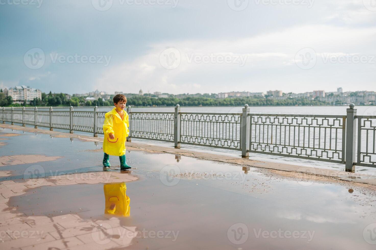 Little boy playing in rainy summer park. Child with umbrella, waterproof coat and boots jumping in puddle and mud in the rain. Kid walking in summer rain Outdoor fun by any weather. happy childhood. photo