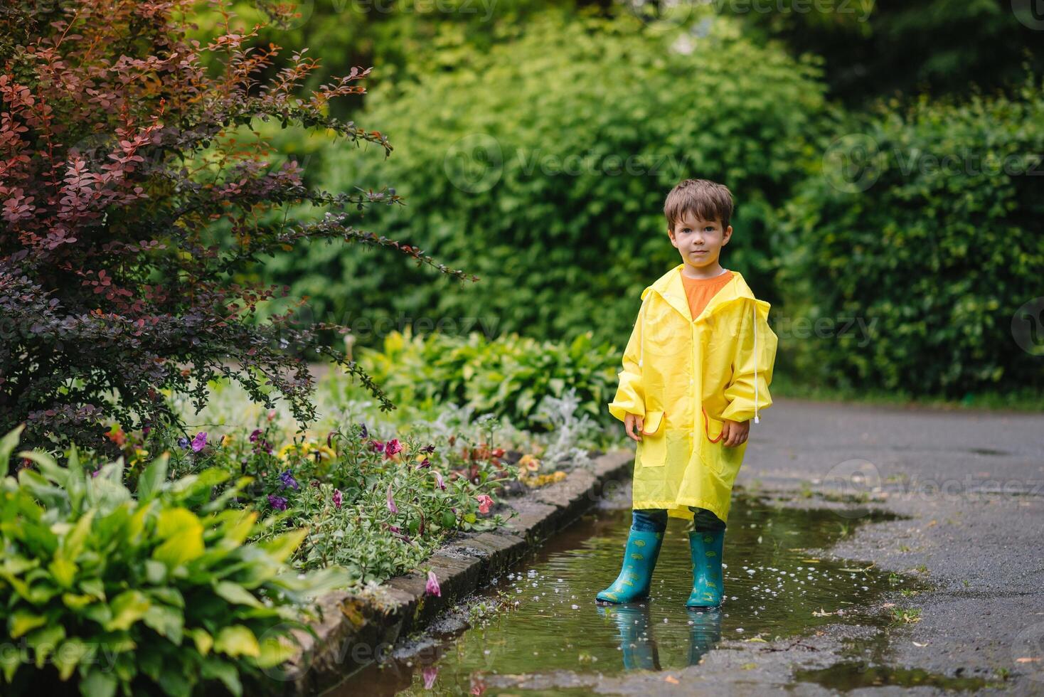 Little boy playing in rainy summer park. Child with umbrella, waterproof coat and boots jumping in puddle and mud in the rain. Kid walking in summer rain Outdoor fun by any weather. happy childhood. photo