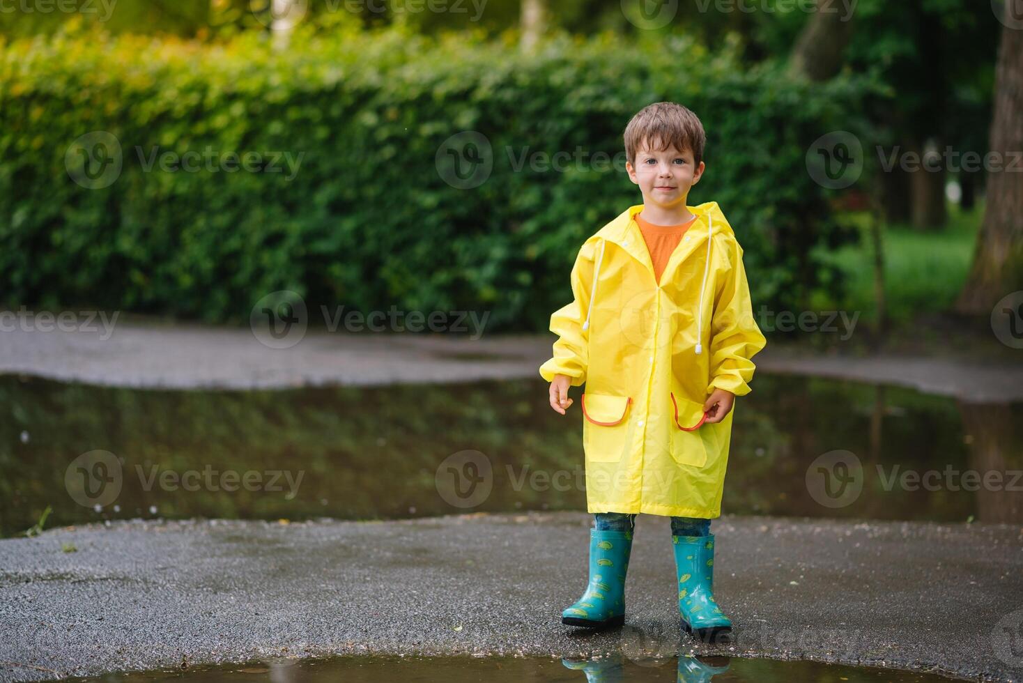 Little boy playing in rainy summer park. Child with umbrella, waterproof coat and boots jumping in puddle and mud in the rain. Kid walking in summer rain Outdoor fun by any weather. happy childhood photo