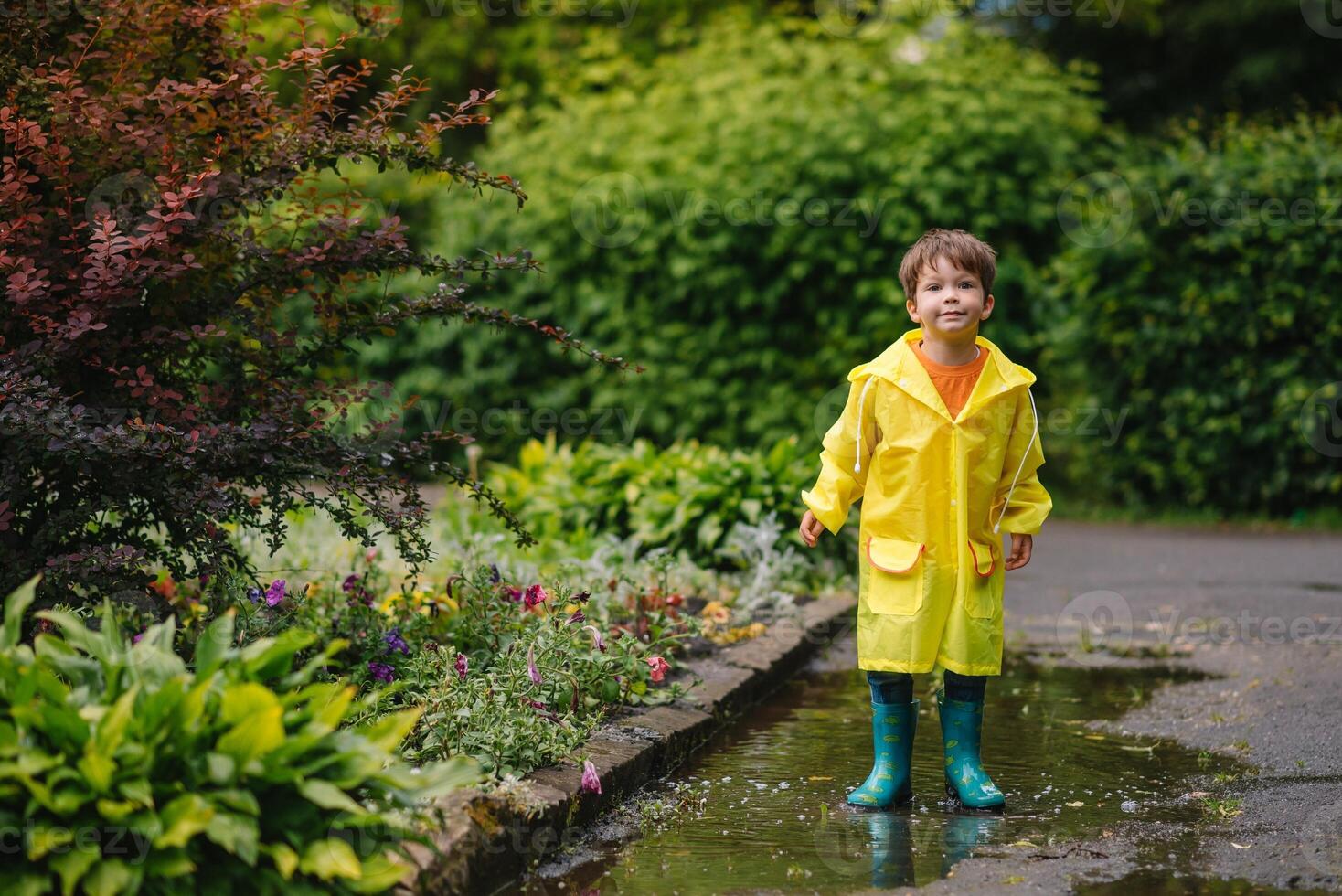 Little boy playing in rainy summer park. Child with umbrella, waterproof coat and boots jumping in puddle and mud in the rain. Kid walking in summer rain Outdoor fun by any weather. happy childhood. photo