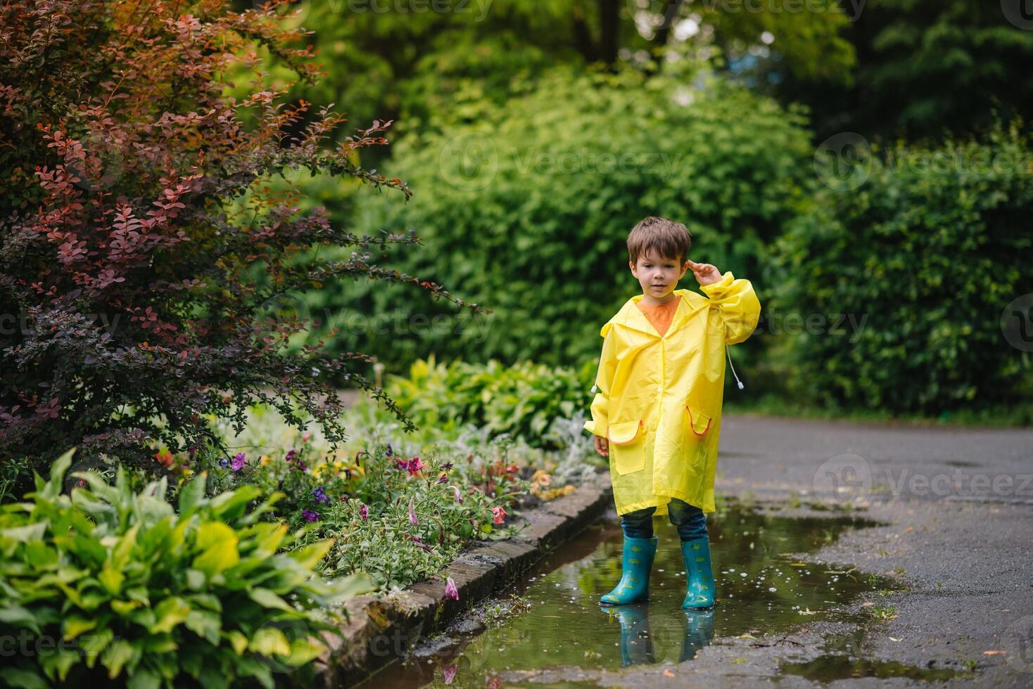 Little boy playing in rainy summer park. Child with umbrella, waterproof coat and boots jumping in puddle and mud in the rain. Kid walking in summer rain Outdoor fun by any weather. happy childhood. photo