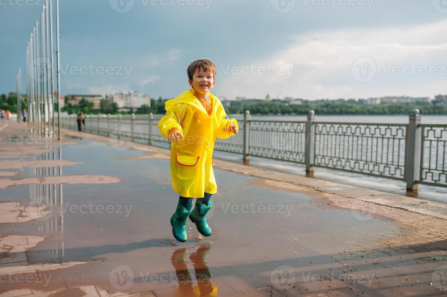 Little boy playing in rainy summer park. Child with umbrella, waterproof coat and boots jumping in puddle and mud in the rain. Kid walking in summer rain Outdoor fun by any weather. happy childhood. photo