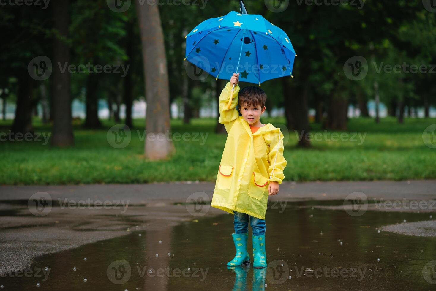 Little boy playing in rainy summer park. Child with umbrella, waterproof coat and boots jumping in puddle and mud in the rain. Kid walking in summer rain Outdoor fun by any weather. happy childhood. photo