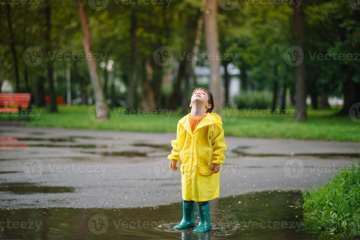Little boy playing in rainy summer park. Child with umbrella, waterproof coat and boots jumping in puddle and mud in the rain. Kid walking in summer rain Outdoor fun by any weather. happy childhood photo