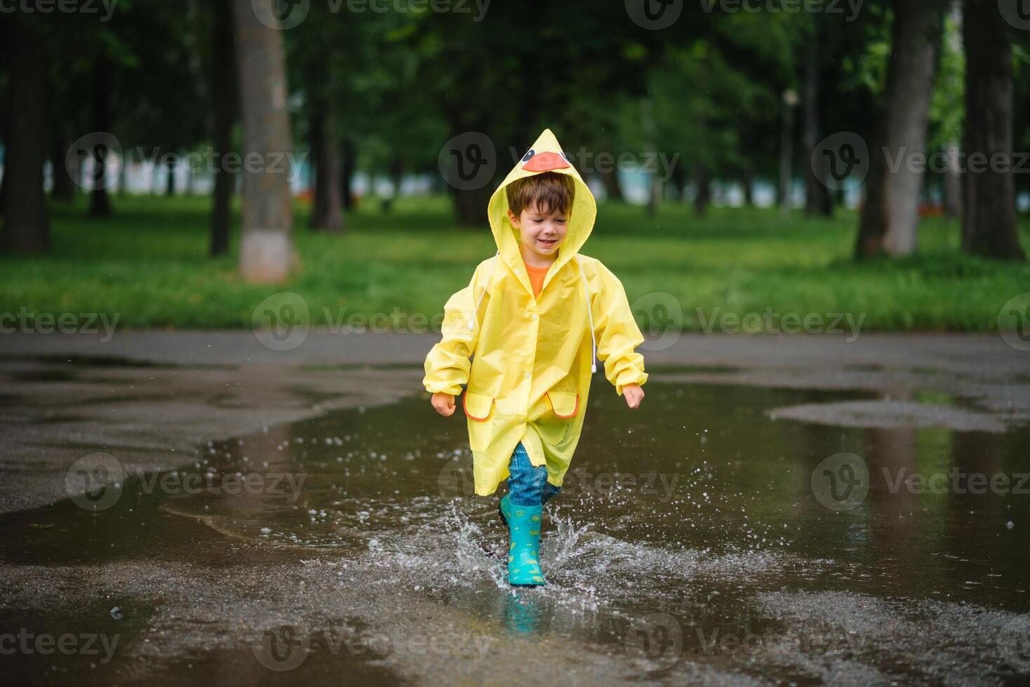 Little boy playing in rainy summer park. Child with umbrella, waterproof coat and boots jumping in puddle and mud in the rain. Kid walking in summer rain Outdoor fun by any weather. happy childhood. photo