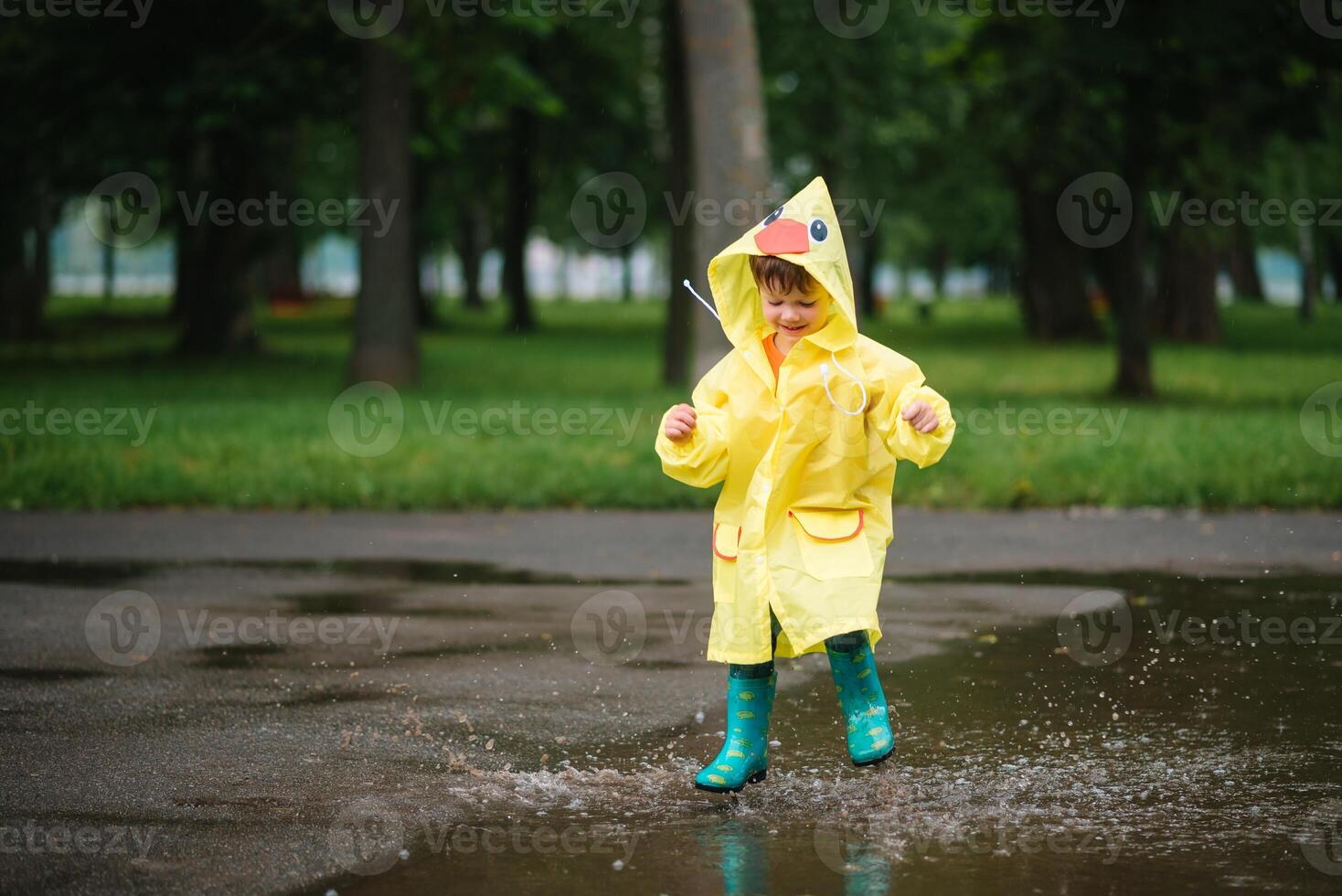 Little boy playing in rainy summer park. Child with umbrella, waterproof coat and boots jumping in puddle and mud in the rain. Kid walking in summer rain Outdoor fun by any weather. happy childhood. photo