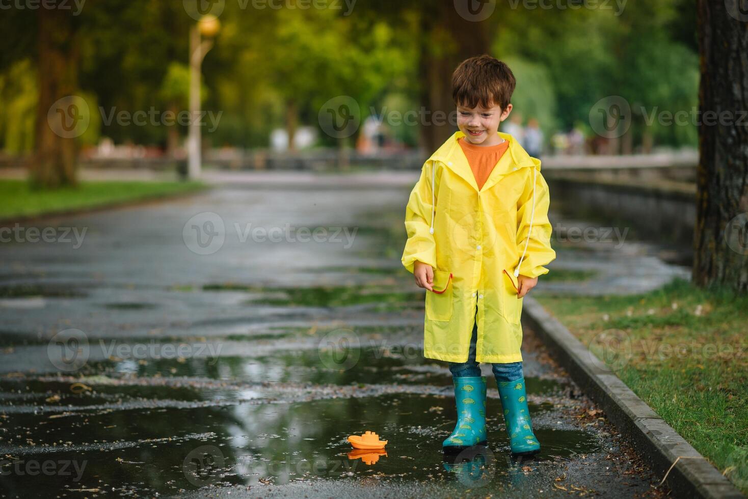 Child playing with toy boat in puddle. Kid play outdoor by rain. Fall rainy weather outdoors activity for young children. Kid jumping in muddy puddles. Waterproof jacket and boots for baby. childhood photo