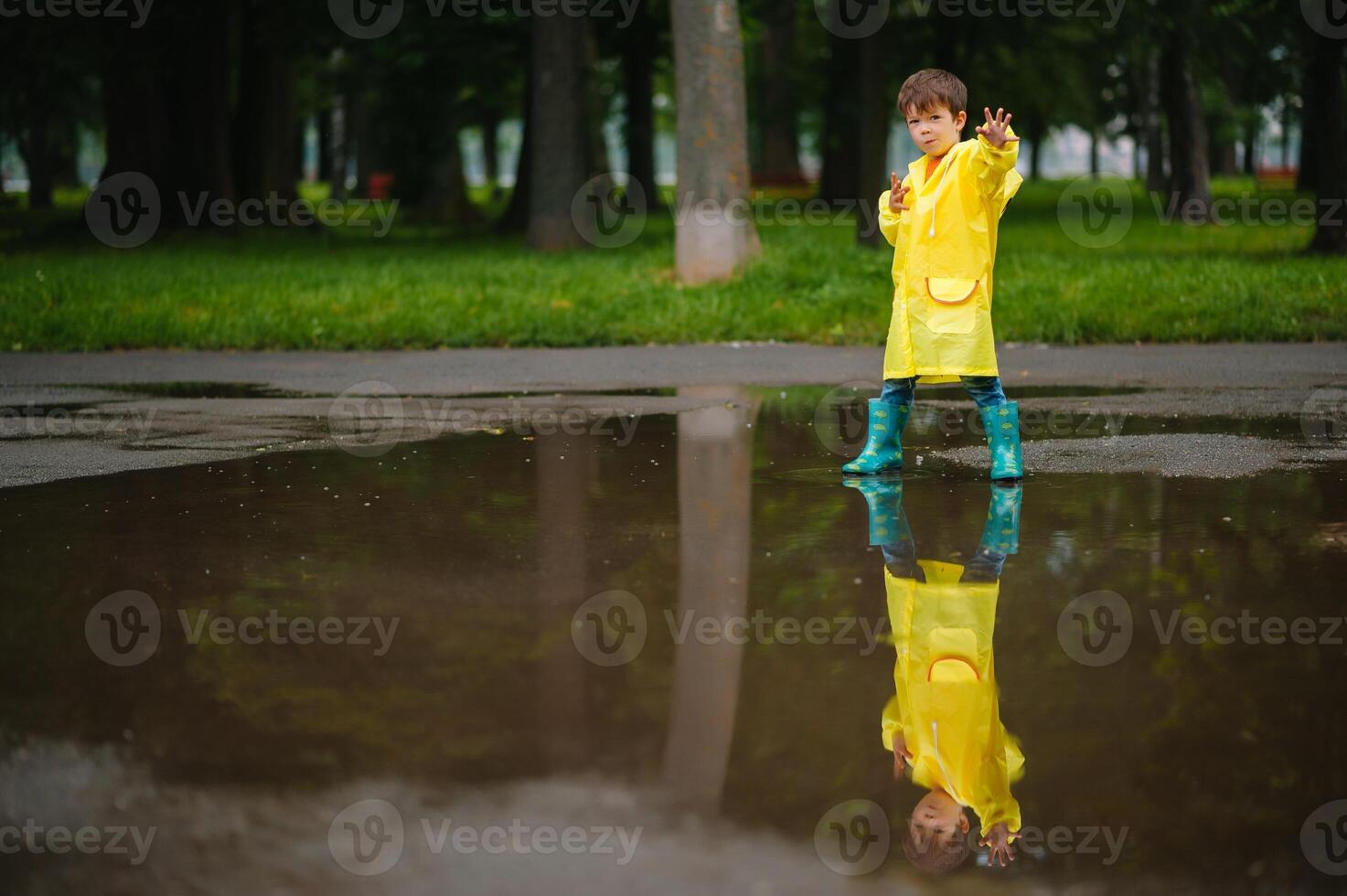 Little boy playing in rainy summer park. Child with umbrella, waterproof coat and boots jumping in puddle and mud in the rain. Kid walking in summer rain Outdoor fun by any weather. happy childhood. photo