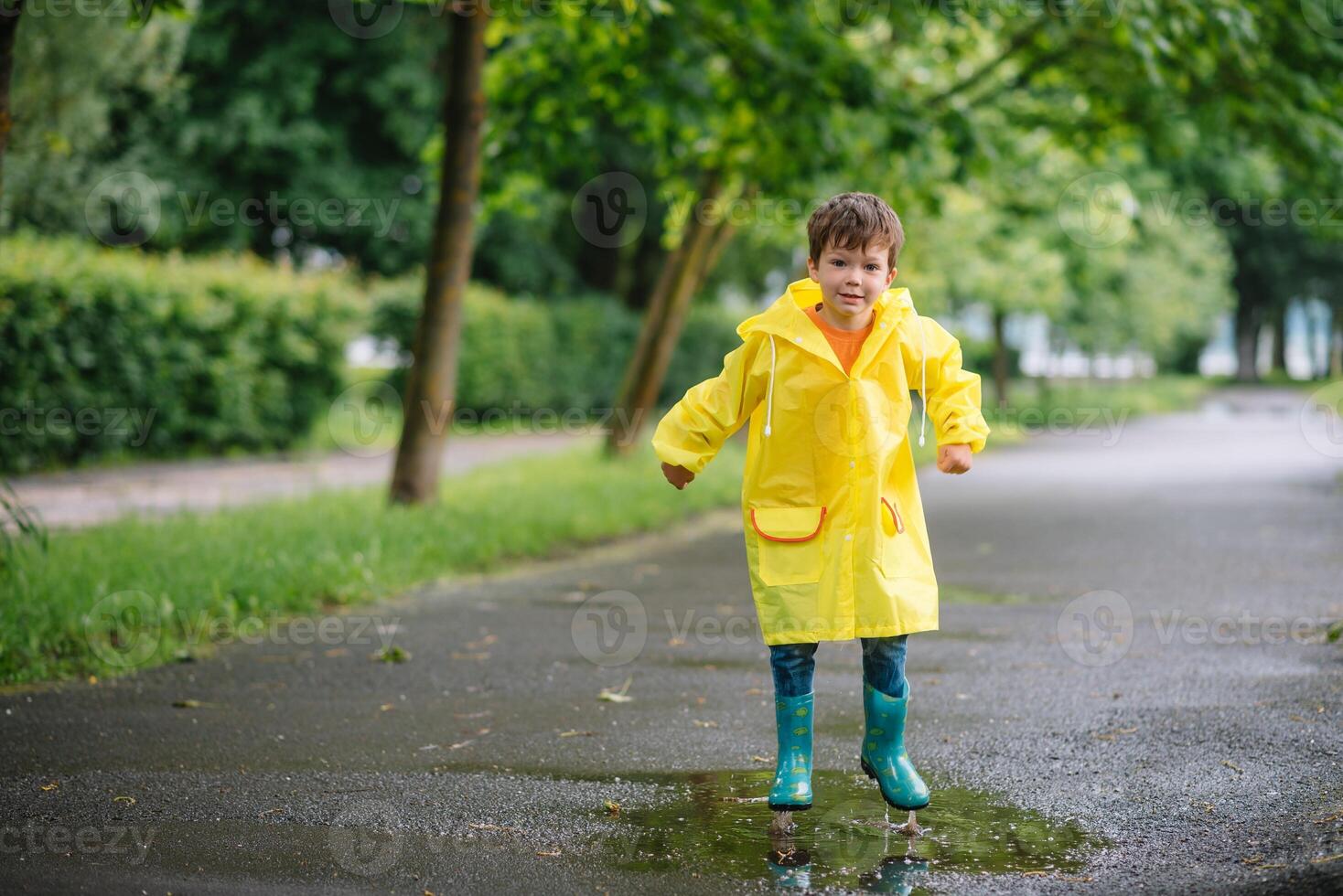 Little boy playing in rainy summer park. Child with umbrella, waterproof coat and boots jumping in puddle and mud in the rain. Kid walking in summer rain Outdoor fun by any weather. happy childhood photo
