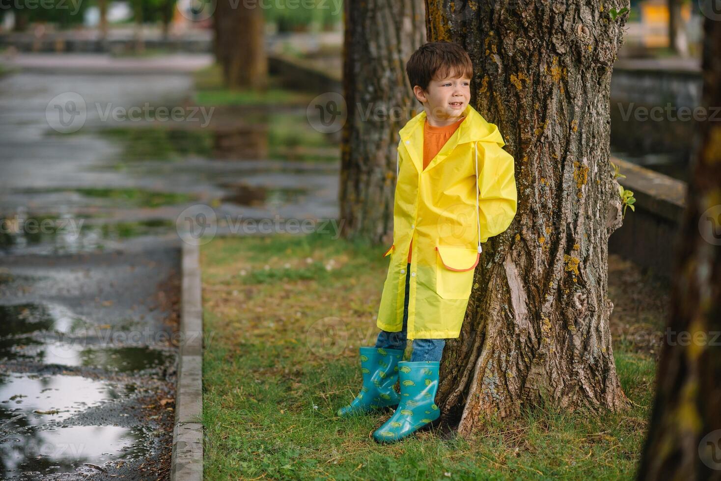 Little boy playing in rainy summer park. Child with umbrella, waterproof coat and boots jumping in puddle and mud in the rain. Kid walking in summer rain Outdoor fun by any weather. happy childhood. photo