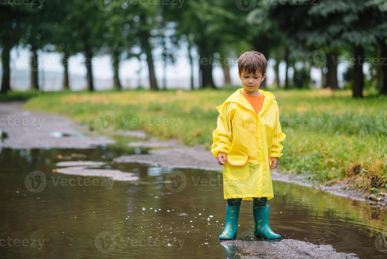 Little boy playing in rainy summer park. Child with umbrella, waterproof coat and boots jumping in puddle and mud in the rain. Kid walking in summer rain Outdoor fun by any weather. happy childhood photo