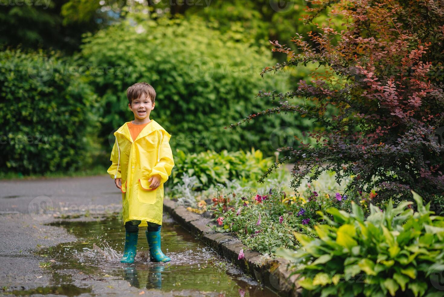 Little boy playing in rainy summer park. Child with umbrella, waterproof coat and boots jumping in puddle and mud in the rain. Kid walking in summer rain Outdoor fun by any weather. happy childhood. photo