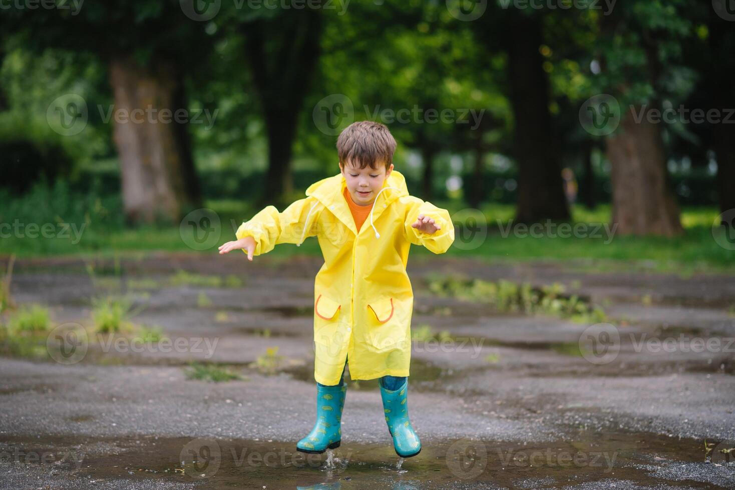 Little boy playing in rainy summer park. Child with umbrella, waterproof coat and boots jumping in puddle and mud in the rain. Kid walking in summer rain Outdoor fun by any weather. happy childhood photo