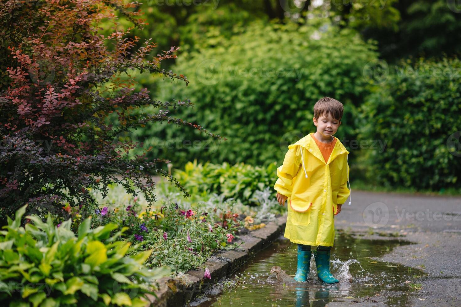 Little boy playing in rainy summer park. Child with umbrella, waterproof coat and boots jumping in puddle and mud in the rain. Kid walking in summer rain Outdoor fun by any weather. happy childhood. photo