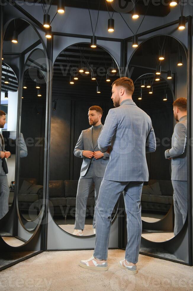 Modern young handsome businessman dressed in classical suit adjusting a tie in front of the mirror while standing in the suit shop photo