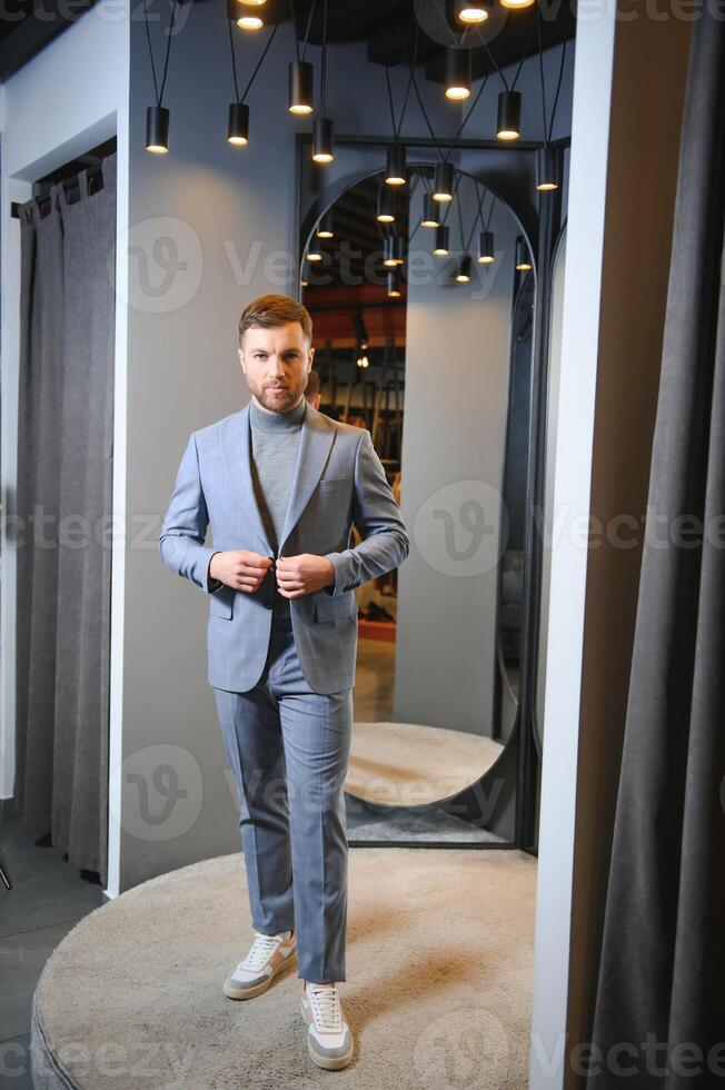 Modern young handsome businessman dressed in classical suit adjusting a tie in front of the mirror while standing in the suit shop photo