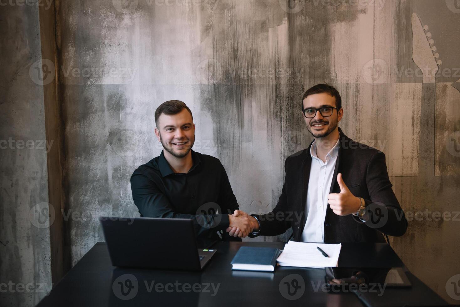 dos empresarios en un oficina sonriente a el cámara mientras trabajando juntos detrás un ordenador portátil computadora. foto