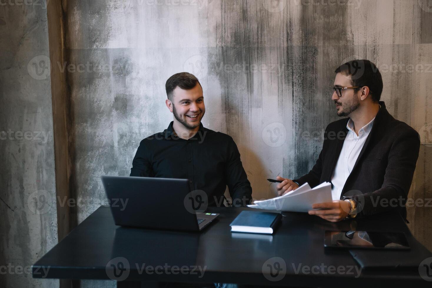 dos empresarios en un oficina sonriente a el cámara mientras trabajando juntos detrás un ordenador portátil computadora. foto