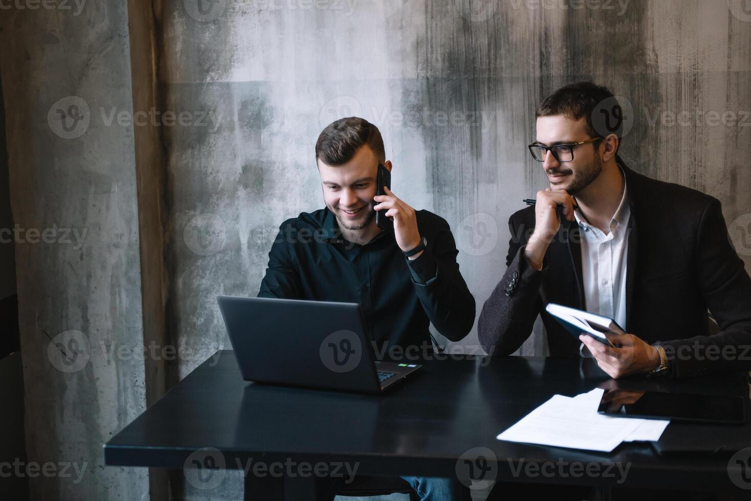dos empresarios en un oficina sonriente a el cámara mientras trabajando juntos detrás un ordenador portátil computadora. foto