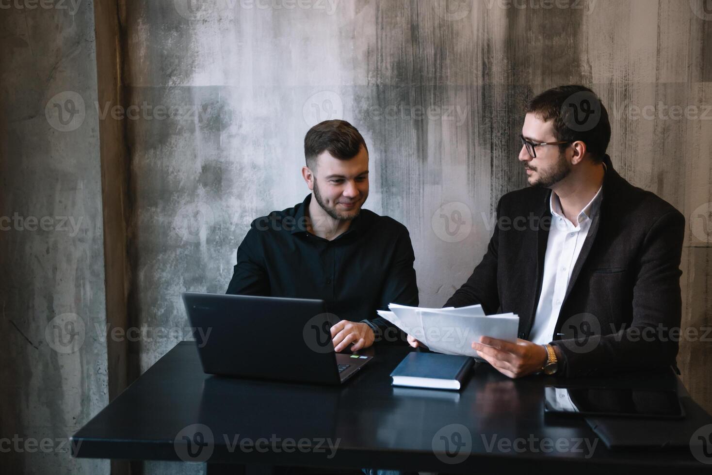 dos empresarios en un oficina sonriente a el cámara mientras trabajando juntos detrás un ordenador portátil computadora. foto