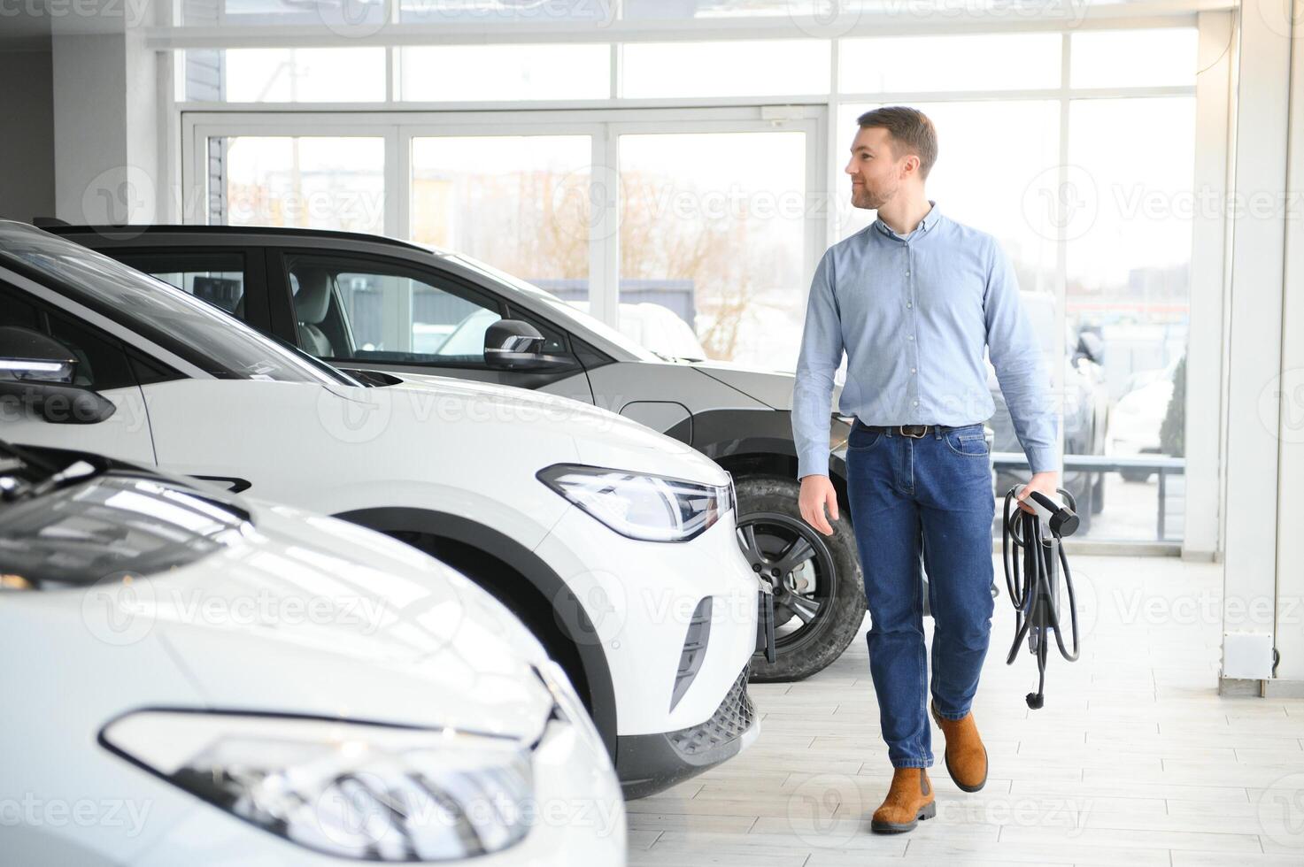Handsome business man holding charging cable for electric car. Caucasian male stands near electric auto in dealership photo