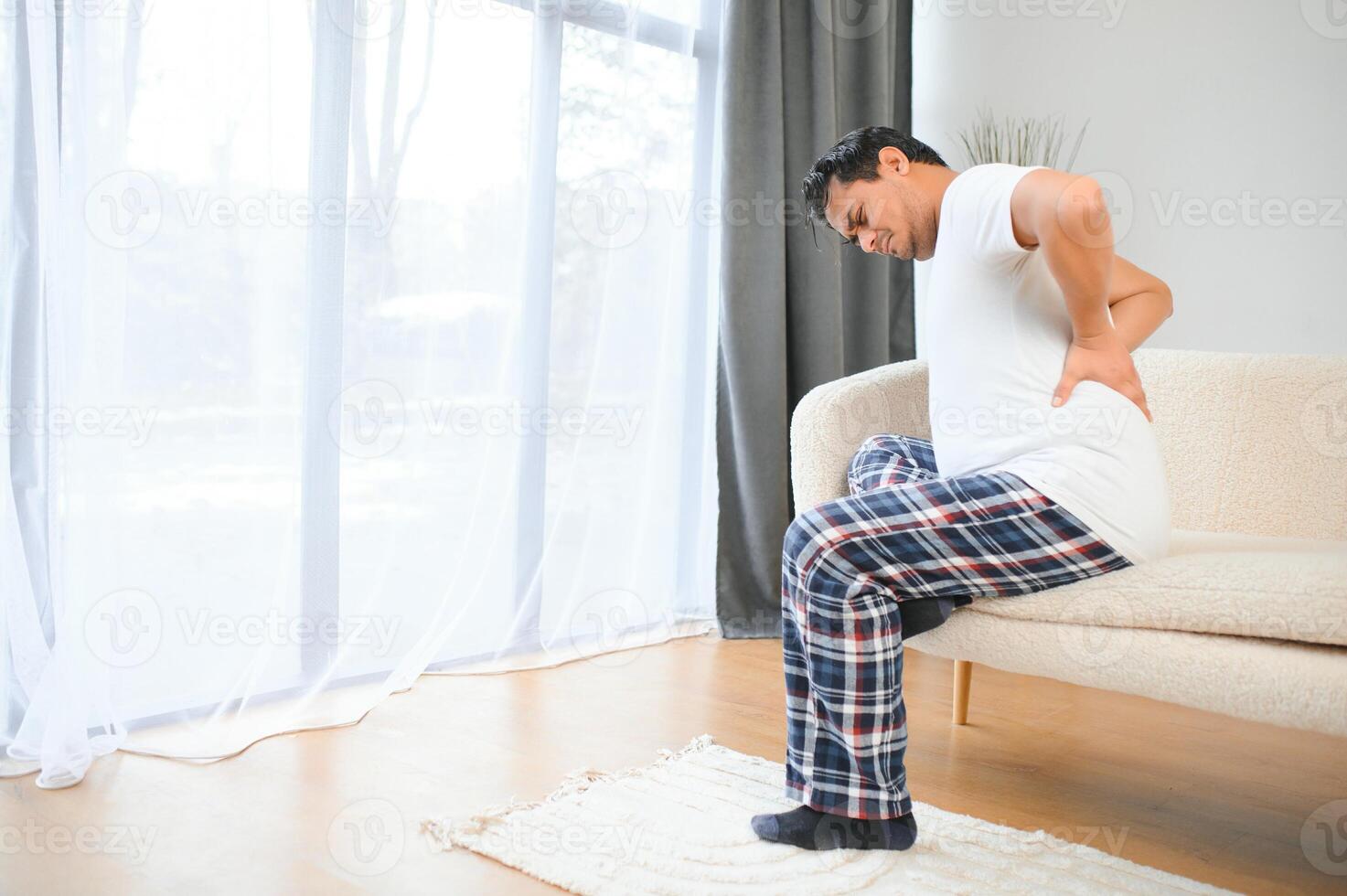 A sick man on Christmas has severe back pain, massages muscle his hand sitting on sofa in living room photo
