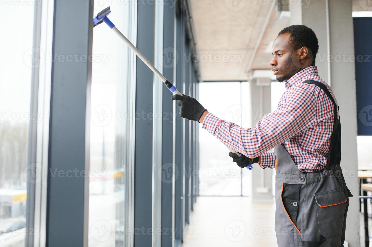 worker of cleaning organization carefully and carefully rubs large window of the office space. A serious African-American in blue overalls wipes the double-glazed window in the office photo
