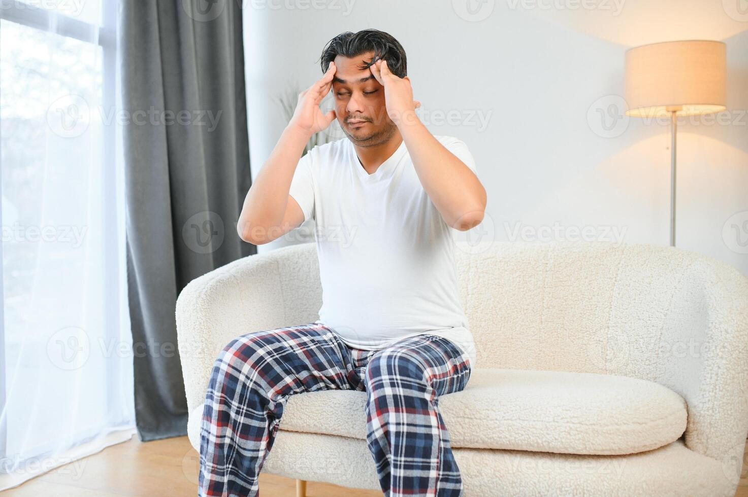 retrato de joven indio chico sufrimiento desde migraña a hogar, oriental hombre sensación indispuesto, conmovedor su templos con cerrado ojos, sufrimiento agudo dolor de cabeza mientras sentado en sofá foto