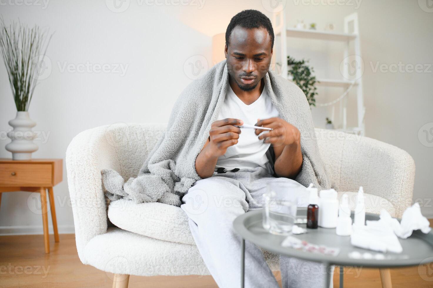Fever. Portrait of sick african american man with thermometer suffering from flu symptoms sitting wrapped in blanket in bed at home photo