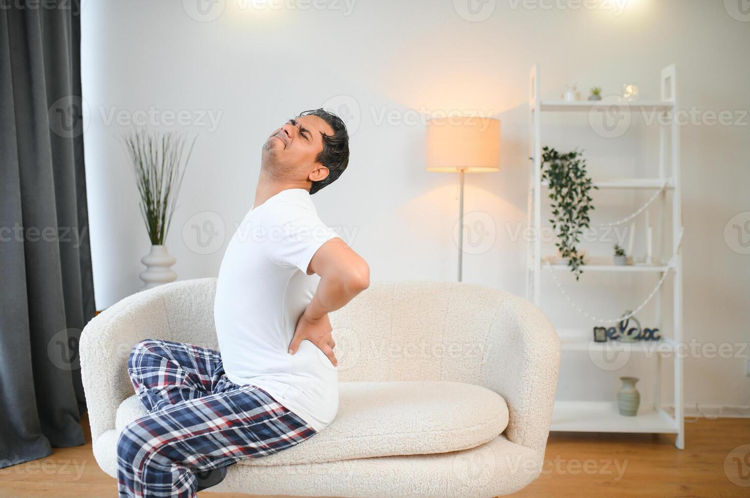 A young Indian man is suffering from severe pain. He sits at home on the sofa and holds his hand behind his back photo