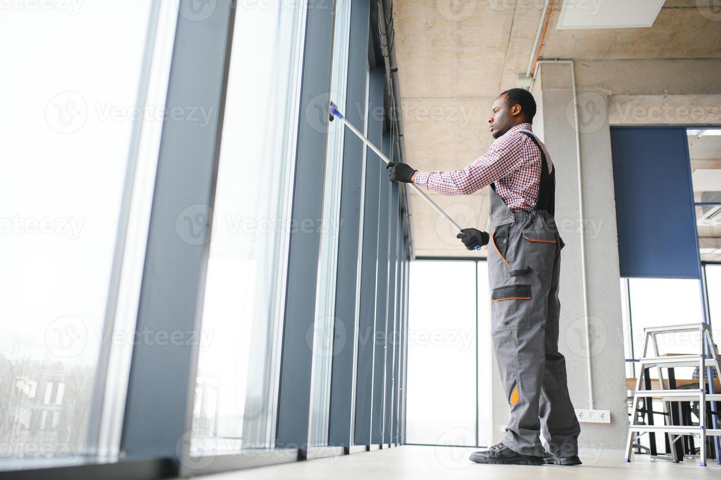 African Male professional cleaning service worker in overalls cleans the windows and shop windows of a store with special equipment photo
