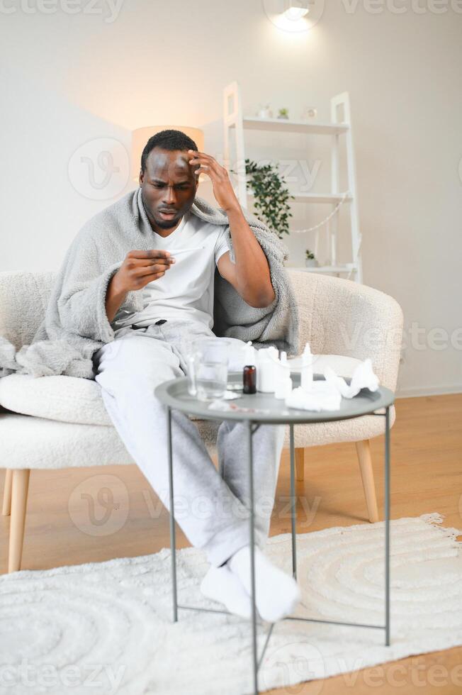 Fever. Portrait of sick african american man with thermometer suffering from flu symptoms sitting wrapped in blanket in bed at home photo