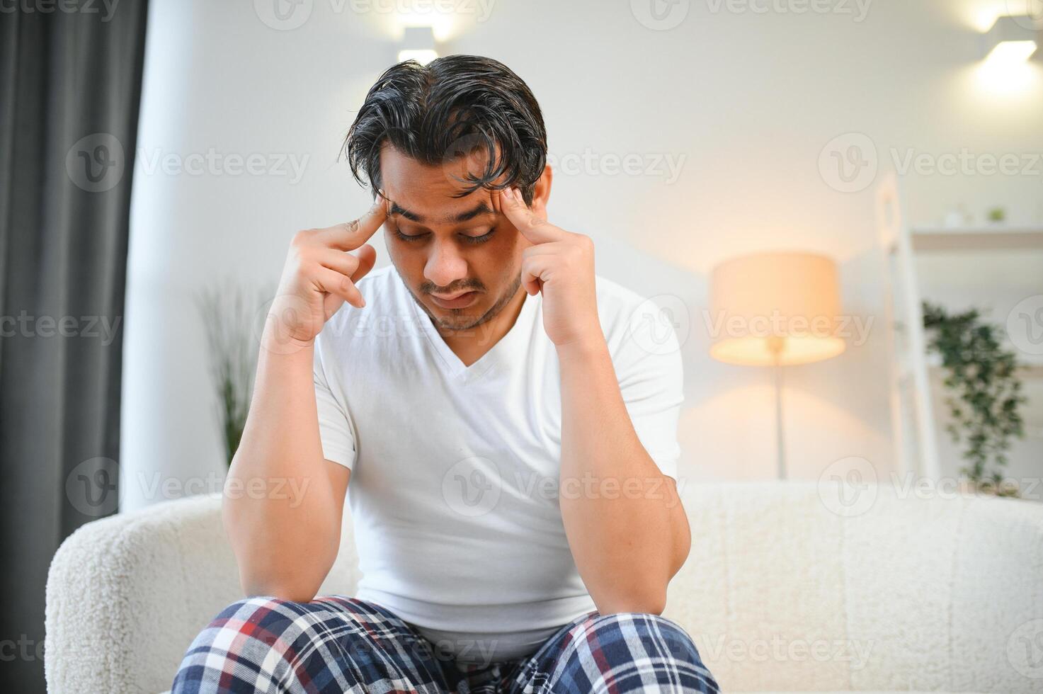 Portrait of young indian guy suffering from migraine at home, eastern man feeling unwell, touching his temples with closed eyes, suffering acute headache while sitting on couch photo