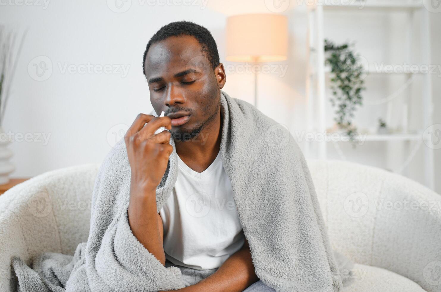 Portrait of a sick young african american man wrapped in a blanket on the sofa. Guy with seasonal flu or cold feel unhealthy with influenza and runny nose at home photo