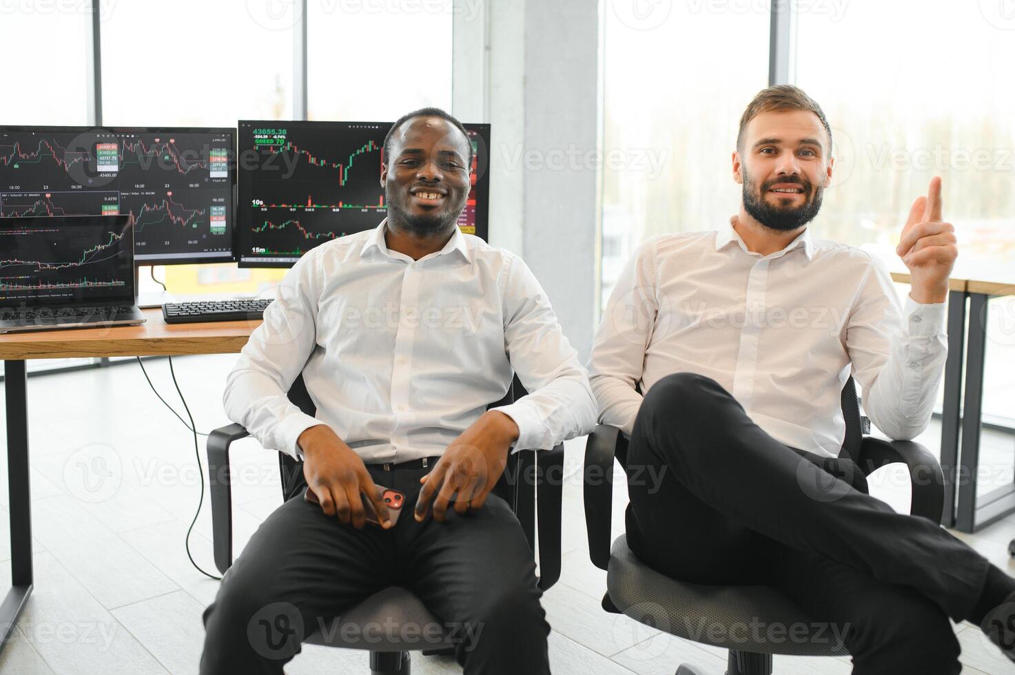 Two diverse colleagues traders talking to each other, sitting in the office in front of multiple computer screens. Stock trading, people, business concept photo