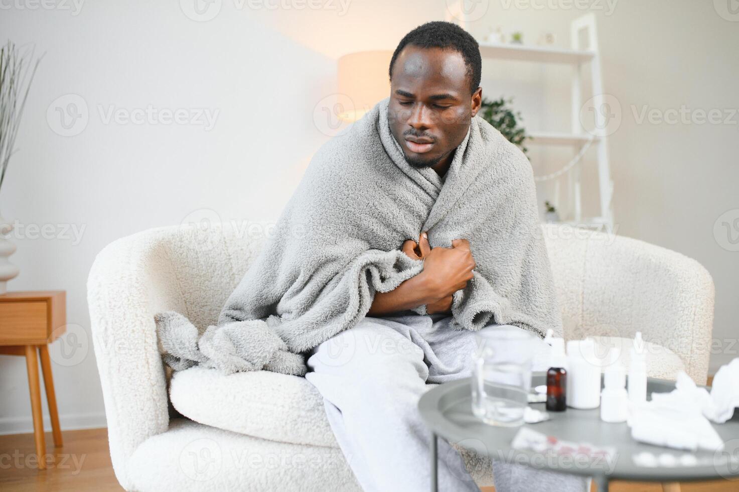 Cold african american young man covered in grey blanket freezing at home, sitting on sofa alone and warming up his hands in living room, copy space photo
