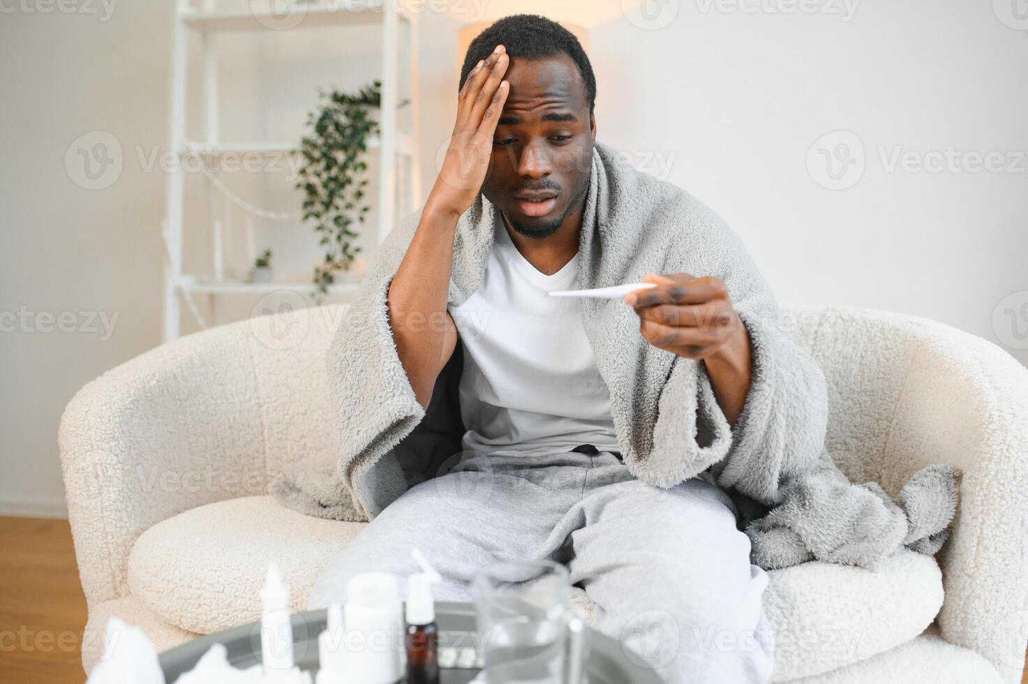 Fever. Portrait of sick african american man with thermometer suffering from flu symptoms sitting wrapped in blanket in bed at home photo