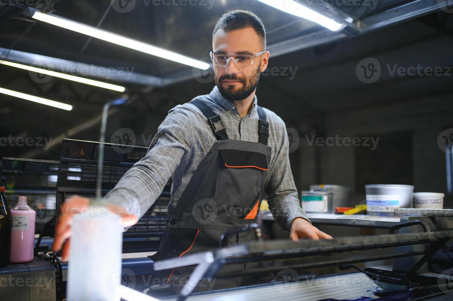 Printing house interior with modern offset print machine and operator in working uniform checking quality and controlling process of print photo