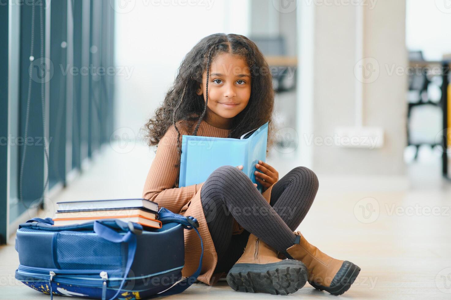 niña sonriente. hermosa africano americano Chica de escuela sonriente mientras sentado cerca ventana y leyendo libro foto