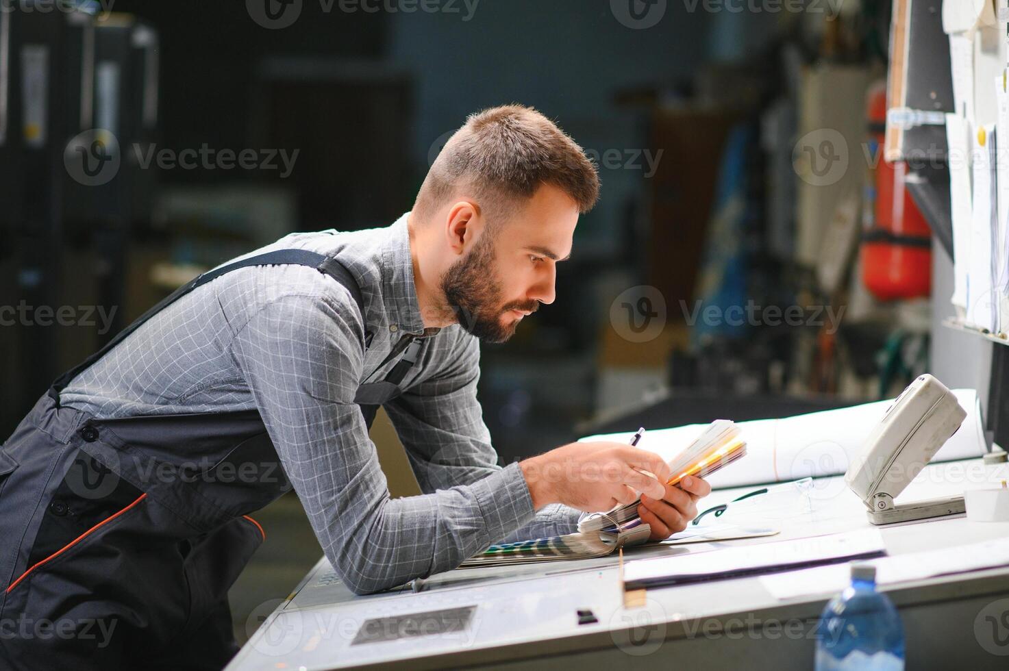 Printing house interior with modern offset print machine and operator in working uniform checking quality and controlling process of print photo